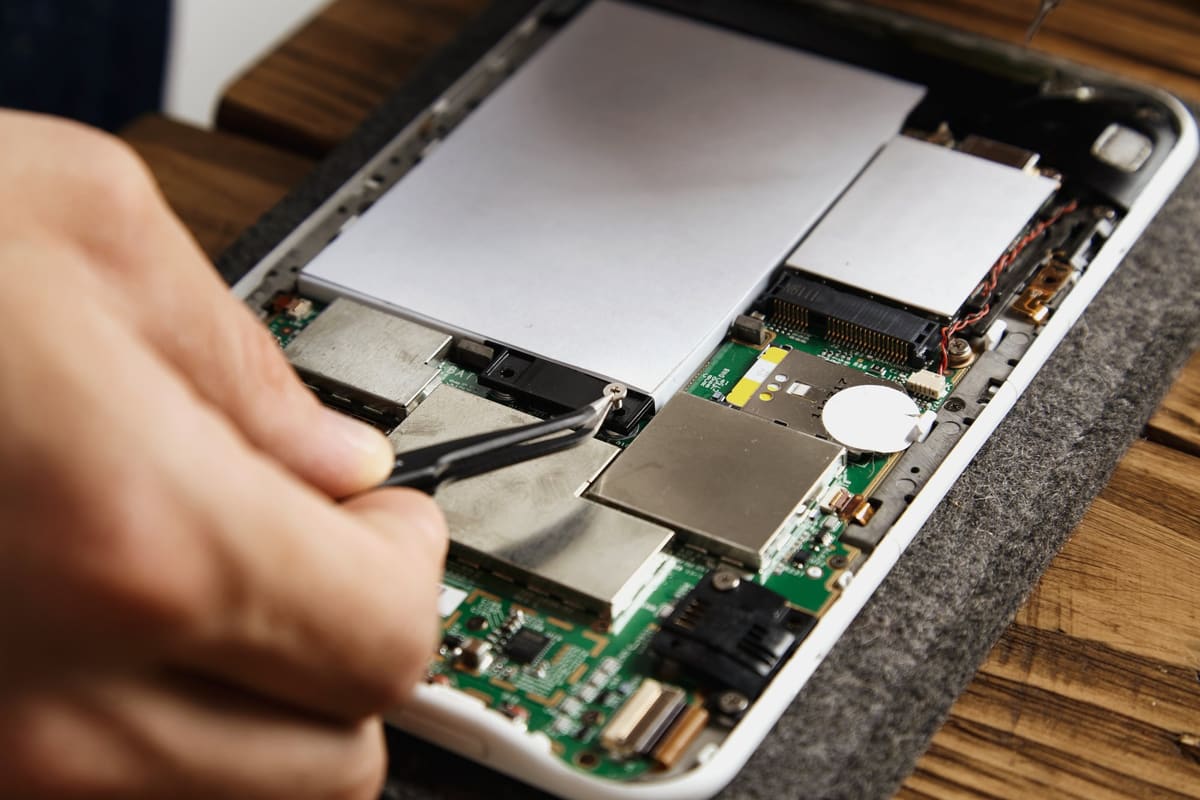 Tweezers removing a small screw near a phone battery on a motherboard